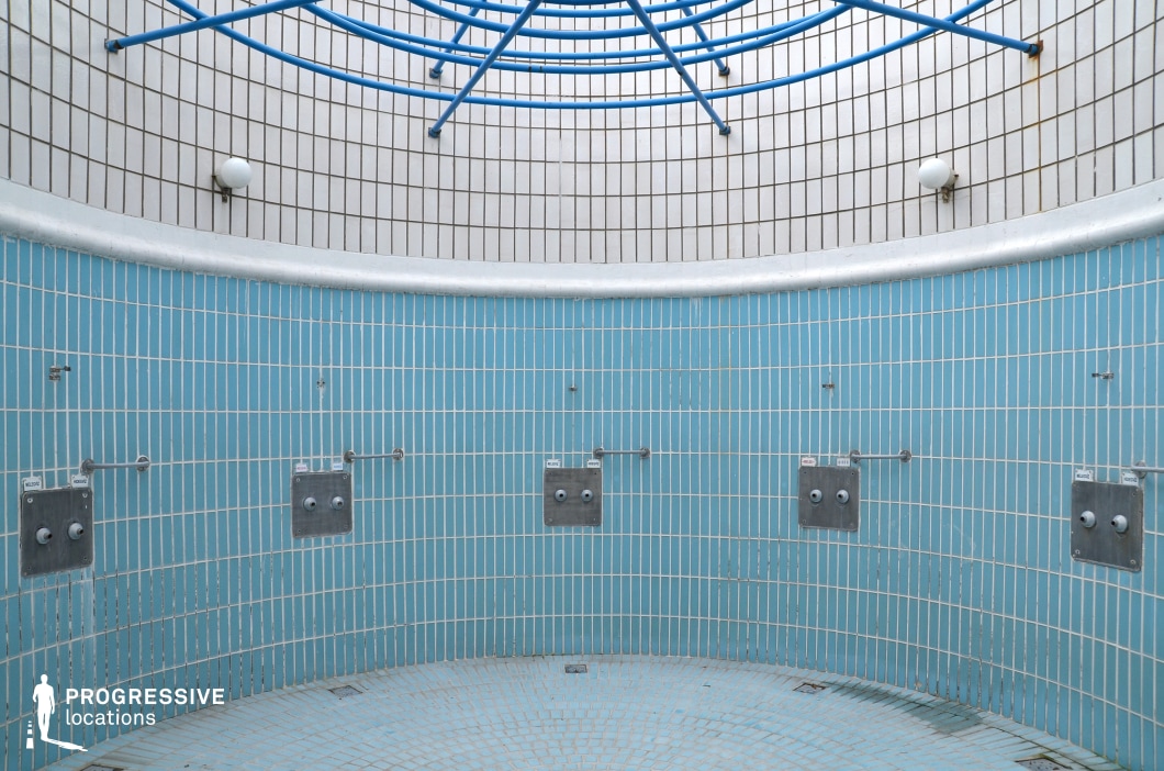 This circular shower facility features a stark, utilitarian design with blue and white vertical tiles covering the curved walls and floor. The industrial-style stainless steel fixtures contrast with the clean tile work, emphasizing a functional and minimalistic aesthetic. Overhead, a geometric blue metal structure adds a subtle architectural element, while evenly spaced wall-mounted light fixtures provide illumination. The enclosed yet open layout creates a unique, almost futuristic atmosphere, making this location ideal for filming industrial, dystopian, or minimalist-themed productions.