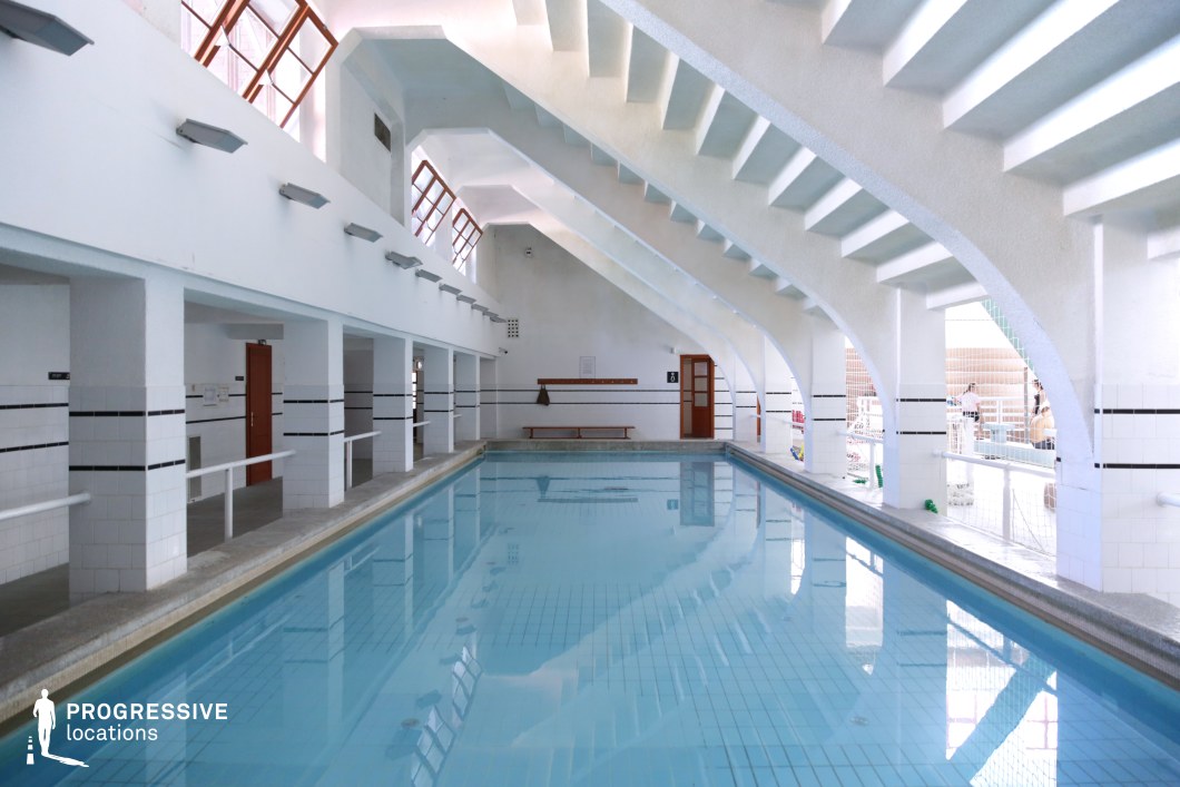 This indoor swimming facility features a striking geometric ceiling with angular, repetitive white beams that create a dynamic architectural pattern. The long, narrow pool is lined with white tiles accented by black stripes, giving it a clean and minimalist aesthetic. Large, slanted windows on the upper walls allow natural light to stream in, enhancing the bright, reflective quality of the water. A row of changing room doors and a simple wooden bench contribute to the functional and utilitarian design. The space is ideal for scenes requiring a vintage or retro-modern sports setting, a serene training environment, or a minimalist aesthetic.