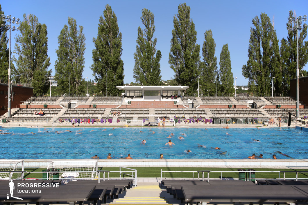 This large outdoor water polo stadium features a competition-size pool surrounded by tiered grandstands constructed from concrete and brick, offering ample spectator seating. The symmetrical layout is enhanced by towering poplar trees, adding a natural backdrop to the sports facility. A central press box overlooks the pool, reinforcing the professional athletic atmosphere. The combination of structured seating, floodlights, and a regulation-size playing area makes this location ideal for filming sports documentaries, training sequences, and high-intensity competition scenes.