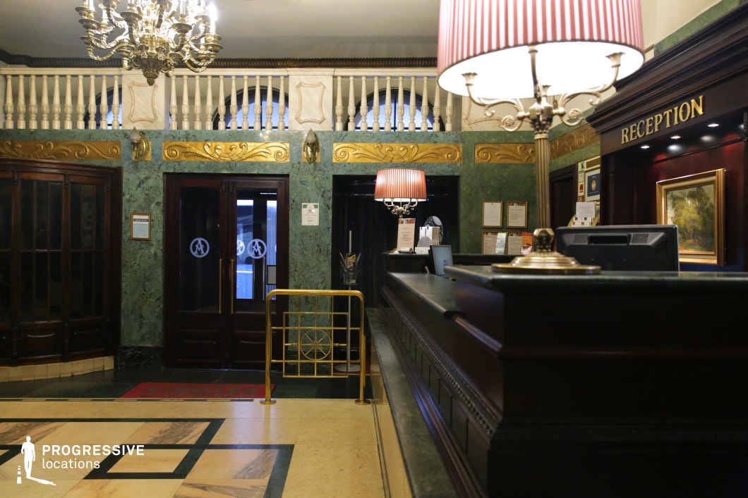 This classic hotel reception area exudes old-world sophistication with its dark wood paneling, deep green marble walls, and ornate gold detailing. A grand chandelier and vintage-style lamps cast a warm glow, highlighting the rich textures and polished surfaces. The symmetrical layout, with an upper-level balcony framed by white balustrades, enhances the sense of grandeur. The refined ambiance and historical character make this an ideal setting for period dramas, mystery thrillers, or noir-inspired films, providing a visually compelling backdrop for intrigue, luxury, and high-stakes interactions.