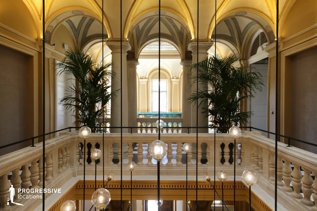 This grand neoclassical atrium features soaring vaulted arches, intricately detailed columns, and a refined color palette of soft gold and cream. A central balustrade overlooks the lower level, while modern hanging globe lights add a contemporary contrast to the historic architecture. Large windows allow natural light to highlight the decorative ceiling details and create a warm, inviting atmosphere. The addition of lush greenery enhances the space’s luxurious ambiance. This location is ideal for historical dramas, sophisticated period films, or stylish thrillers, offering a cinematic backdrop that balances classical grandeur with a modern aesthetic.