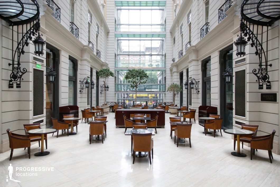 This luxurious atrium lounge showcases a sophisticated blend of classical architecture and contemporary glass design. The symmetrical space features high white walls adorned with intricate black wrought-iron lanterns and decorative balcony railings, creating an air of historic grandeur. A glass ceiling and full-height windows flood the space with natural light, offering a seamless connection between indoor and outdoor environments. Warm-toned leather chairs and dark wooden tables provide an inviting seating arrangement, ideal for upscale dining or social gatherings. This refined setting is perfect for high-end productions, luxury branding, or architectural showcases.