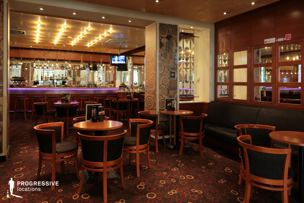 This inviting bar and lounge area features a blend of modern and classic elements, creating a cozy yet sophisticated atmosphere. The space is defined by warm wooden furnishings, a patterned carpet with circular motifs, and an illuminated bar with mirrored panels reflecting ambient lighting. A ceiling adorned with embedded lights enhances the inviting mood, while decorative wall panels add a touch of artistic flair. The seating arrangement includes plush black cushioned benches and wooden chairs, providing a comfortable and social environment. Ideal for nightlife scenes, upscale bar settings, or relaxed social gatherings in film and photography productions.