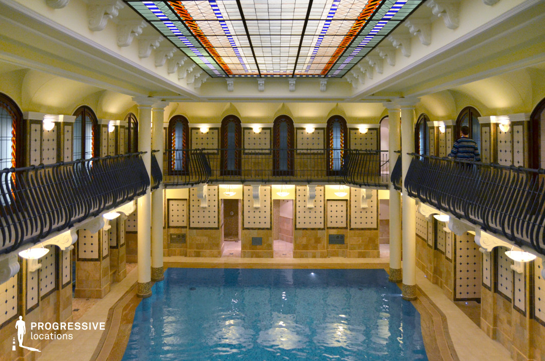 This opulent indoor pool features classical architecture with elegant columns, arched windows, and intricate tiled walls. The central pool reflects the warm lighting, creating a tranquil and luxurious ambiance. A striking stained glass ceiling adds a touch of grandeur, casting soft, colorful light across the space. The wrought-iron railings on the upper level enhance the historical charm, making this an ideal setting for period dramas, high-end spa scenes, or a refined relaxation retreat.