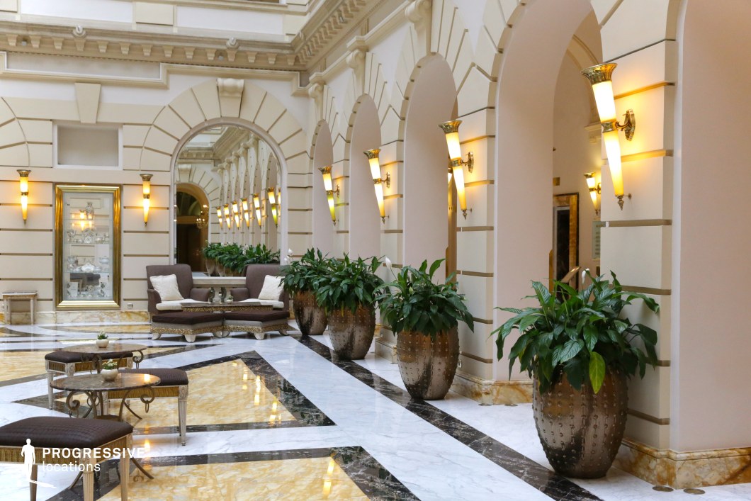 This sophisticated hotel lounge features a grand neoclassical design with arched colonnades, intricate wall moldings, and luxurious marble flooring. The black, white, and gold-veined marble patterns enhance the sense of opulence, while warm lighting fixtures create an inviting atmosphere. Plush seating arrangements with decorative cushions and ornate side tables add a refined touch, complemented by lush greenery in metallic planters. The space is ideal for high-end film productions, luxury brand promotions, or scenes requiring a regal and timeless backdrop.