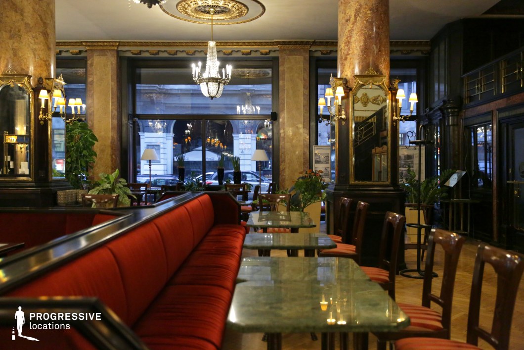 This refined café setting combines rich classical elegance with a warm, intimate ambiance. The plush red velvet seating, polished green marble tabletops, and dark wood paneling create a striking contrast, while ornate chandeliers and gilded mirrors reflect soft, ambient lighting. Grand marble columns and intricate gold detailing further enhance the space’s old-world sophistication. The large windows frame a vibrant cityscape, seamlessly blending the interior’s timeless charm with the urban environment. This location is ideal for period dramas, noir thrillers, or romantic narratives, providing a visually captivating backdrop for refined conversations, intrigue, or secret rendezvous.