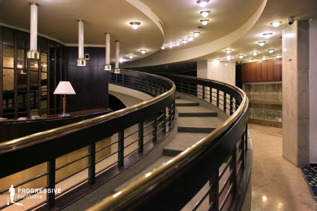 This striking curved staircase embodies mid-century modern aesthetics with dark wood railings, brass accents, and soft ambient lighting. The continuous flow of the structure, combined with rounded ceiling panels lined with recessed lights, creates a sense of movement and sophistication. The surrounding area features wood-paneled walls, reflective surfaces, and minimalist furniture, enhancing the retro-futuristic atmosphere. Ideal for scenes requiring a stylish yet slightly mysterious ambiance, this space lends itself to period dramas, luxury hotel settings, or high-end corporate environments.