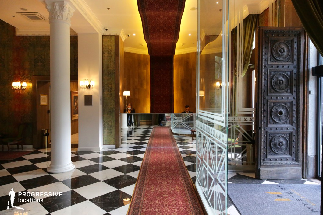 This hotel lobby features a striking blend of classical architecture and modern design elements. The black-and-white checkerboard marble flooring exudes sophistication, complemented by an elongated red Persian-style runner that extends toward the mirrored reception desk. Ornate white columns with intricate capitals frame the space, while antique-style wall sconces cast a warm glow. The grand entrance is defined by a massive carved wooden door, partially open, revealing a glass partition with geometric detailing. The mix of rich green drapery, textured wall finishes, and reflective surfaces creates an opulent yet welcoming ambiance, ideal for luxury-themed productions or high-end fashion shoots.