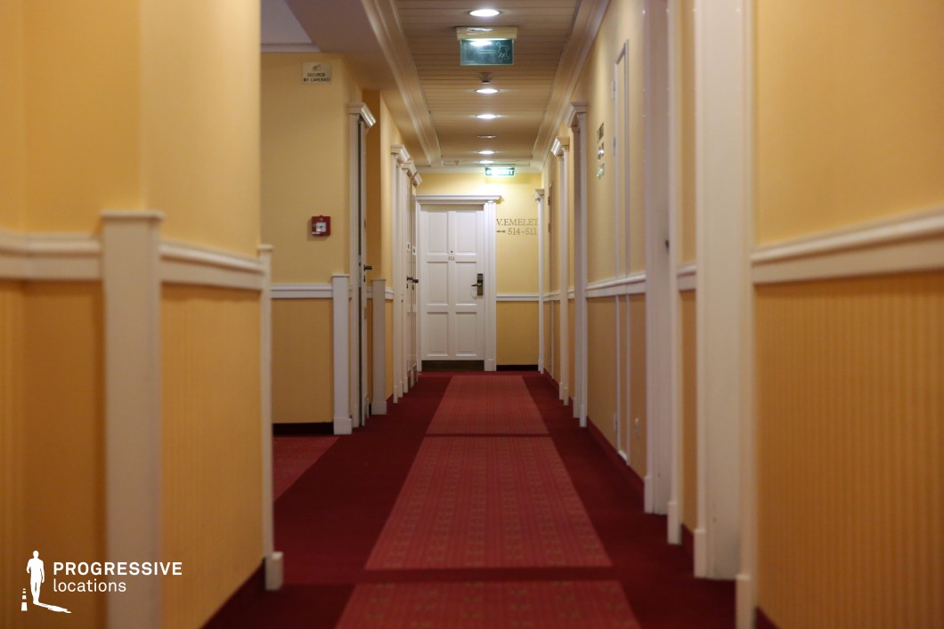 This hotel corridor features a long, symmetrical layout with soft yellow walls accented by white paneling and moldings, creating a refined yet welcoming atmosphere. The deep red carpet with a subtle pattern extends toward a central white door, enhancing the sense of depth and perspective. Recessed ceiling lights provide even illumination, while small architectural details, such as framed doors and decorative wall elements, add to the traditional aesthetic. The balanced composition and warm color palette make this location ideal for filming drama, period films, or mystery scenes requiring an upscale yet slightly enigmatic setting.