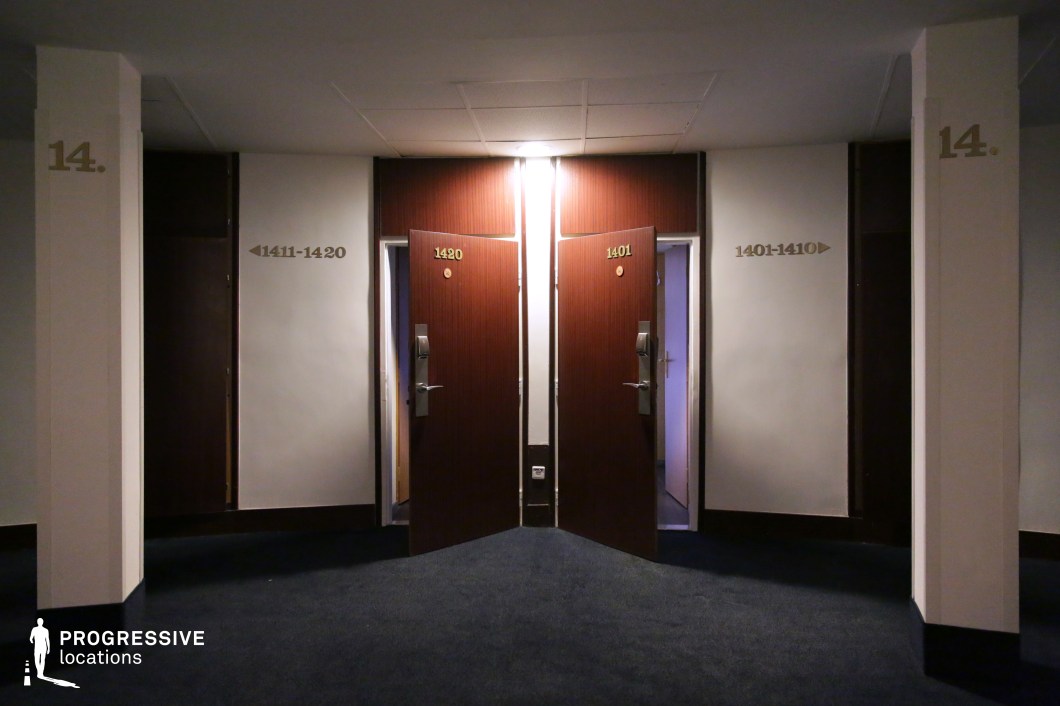 This hotel corridor features a symmetrical design with two slightly ajar wooden doors, each framed by dark wood paneling and illuminated by a single overhead light. The deep blue carpet contrasts with the neutral walls, which are marked with bold room numbers and directional signage. The dim lighting and sharp angles create a moody, enclosed atmosphere, ideal for building suspense. The setting is well-suited for thriller, mystery, or noir films, emphasizing secrecy, tension, or an eerie, enigmatic ambiance.