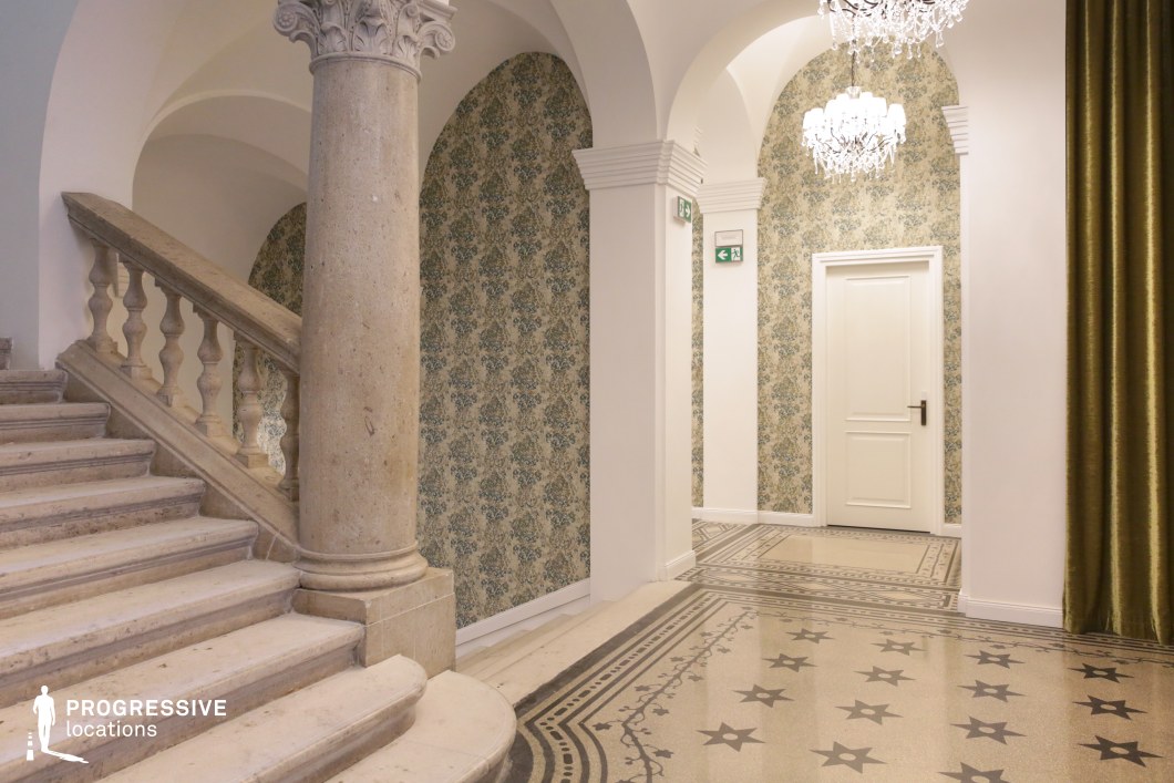 This grand staircase and corridor feature classical architectural elements, including ornate stone columns, arched ceilings, and intricate patterned flooring. The textured wallpaper adds a refined, vintage aesthetic, while crystal chandeliers provide soft, ambient lighting. The elegant curve of the staircase, combined with the symmetry of the archways, creates a visually striking composition, making this space ideal for luxury hotel settings, historical films, or period-piece productions.