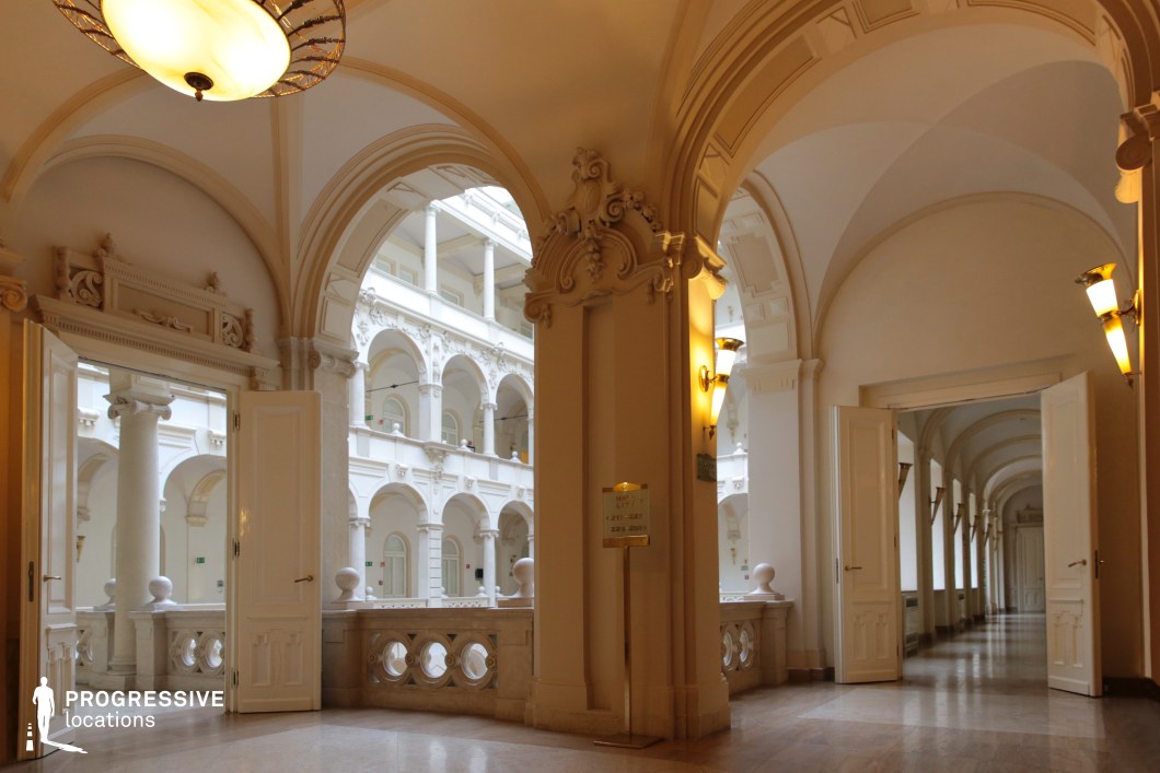 This grand neoclassical corridor is defined by sweeping arches, intricate moldings, and an open view of a central courtyard framed by colonnades. The warm lighting accentuates the detailed stucco work and high ceilings, while the polished stone floors add to the sense of refinement. Open doorways lead into additional hallways, creating an air of mystery and elegance. This location is perfect for historical dramas, aristocratic intrigue, or psychological thrillers, offering a sophisticated setting for moments of revelation, power struggles, or clandestine meetings in a luxurious environment.