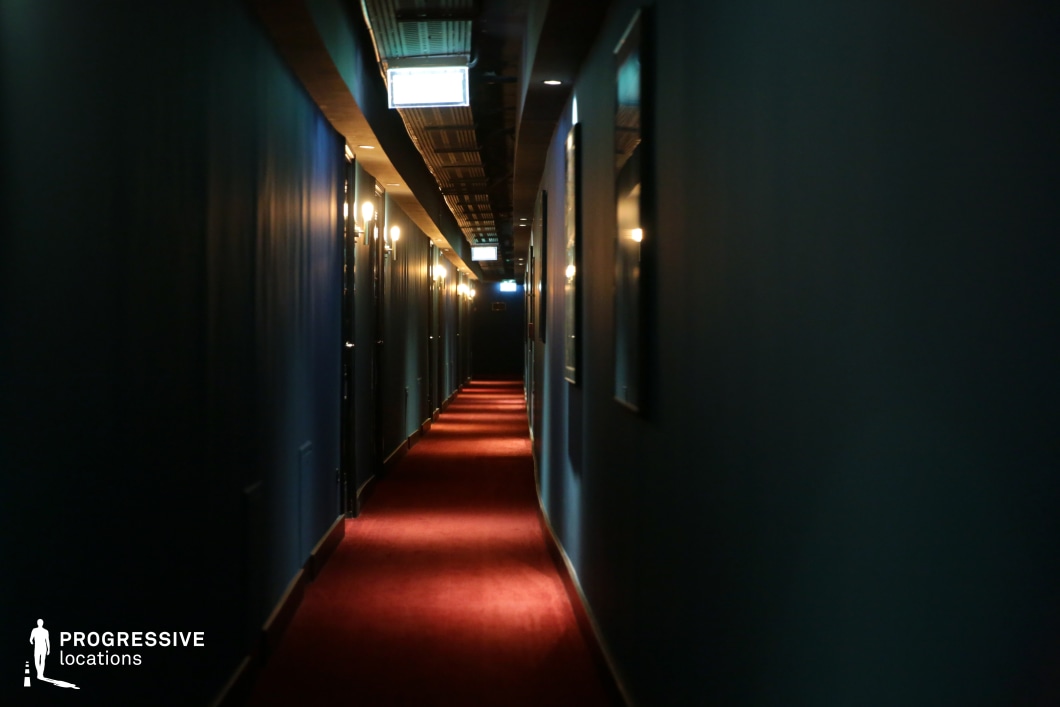 This long, narrow hotel corridor exudes an eerie and atmospheric quality with its deep green walls, moody lighting, and a rich red carpet that adds a dramatic contrast. The ceiling features exposed industrial-style elements, enhancing the sense of depth, while the soft glow from wall sconces creates a rhythmic pattern along the hallway. The vanishing point at the far end intensifies the perspective, making this an ideal filming location for psychological thrillers, horror, or noir-inspired mystery scenes that require suspense and an unsettling ambiance.