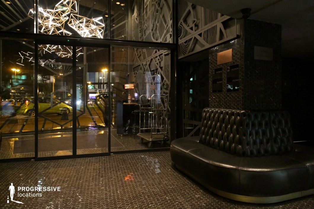 This sophisticated hotel lobby features a bold, futuristic design with dark-toned materials, reflective surfaces, and geometric lighting fixtures. The glossy hexagonal tile flooring and tufted leather seating create a luxurious yet moody atmosphere, while intricate metalwork patterns on the walls and glass doors add depth and texture. The ambient glow from the sculptural chandelier enhances the sleek aesthetic, making this an ideal setting for filming noir thrillers, cyberpunk-inspired stories, or high-end dramas that require a striking, modern backdrop.