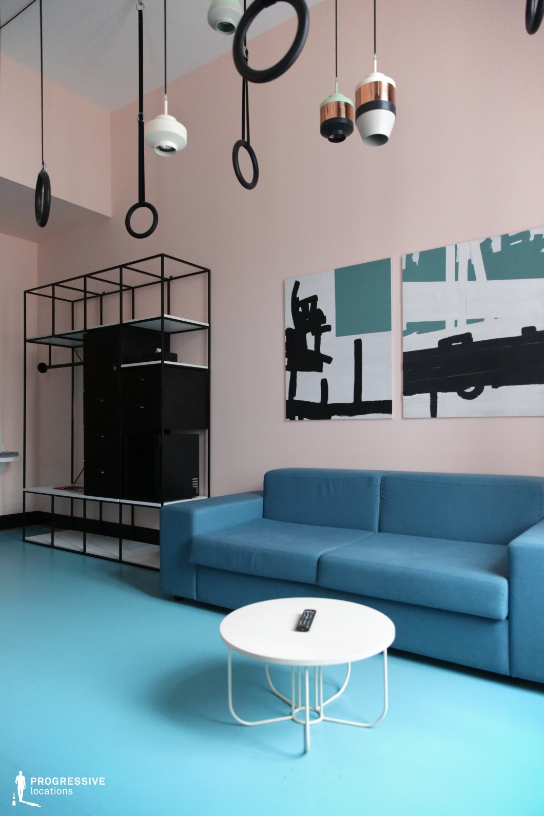 This contemporary living space features a striking combination of pastel pink walls, a vibrant blue floor, and a matching sofa, creating a visually engaging contrast. Black geometric shelving adds structure, while abstract artwork reinforces the modern aesthetic. The ceiling is adorned with unique pendant lights and circular hanging elements, enhancing the artistic and unconventional design. The mix of bold colors, minimalist furniture, and artistic details makes this an ideal filming location for indie films, creative dramas, or futuristic narratives with a stylish and avant-garde setting.