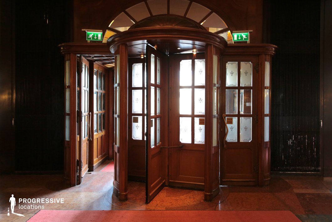This grand entrance features a vintage wooden revolving door with elegant frosted glass panels adorned with delicate motifs, framed by rich wood paneling and arched transom windows. The deep tones of the polished wood contrast with the soft glow of natural light filtering through the glass, creating a refined and timeless aesthetic. The intricate craftsmanship and warm, inviting materials enhance the space’s historic charm. This location is ideal for classic hotel or high-society settings in period dramas, mystery thrillers, or espionage films, providing a sophisticated and cinematic backdrop for moments of arrival, departure, or clandestine exchanges.