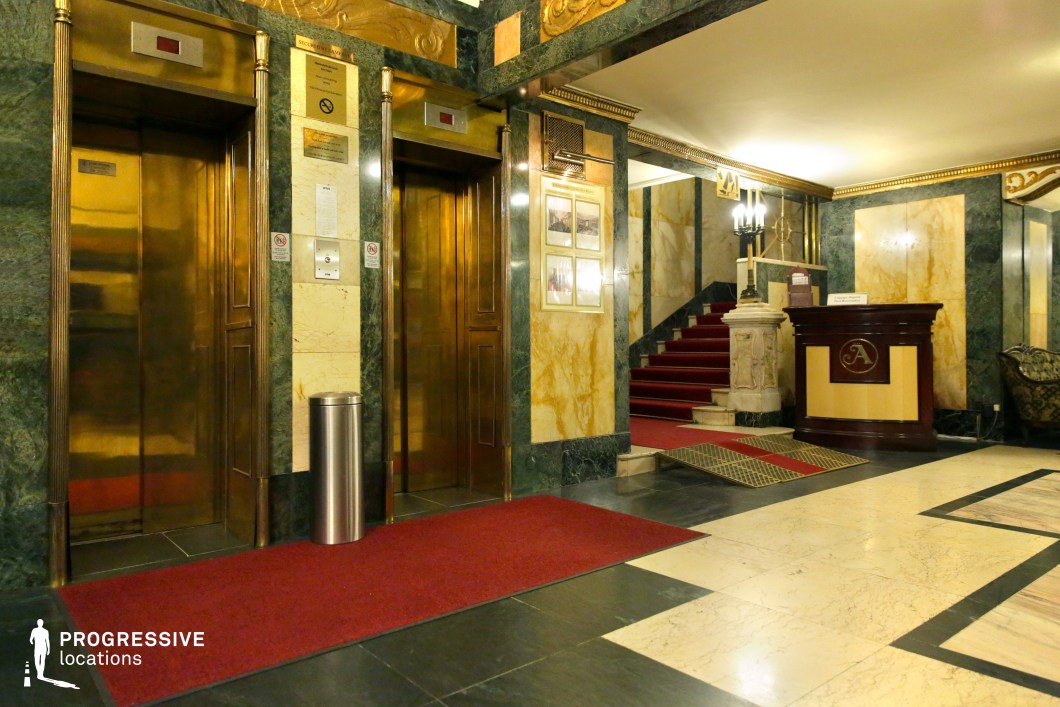This opulent hotel lobby blends Art Deco and classical influences, featuring polished gold-toned elevator doors framed by deep green and beige marble walls with intricate gilded detailing. A plush red carpet leads toward a grand staircase, accentuated by ornate balustrades and warm ambient lighting. The space’s geometric floor patterns and rich materials create a refined yet slightly mysterious atmosphere. This setting is ideal for period dramas, noir thrillers, or psychological suspense films, offering a luxurious yet enigmatic backdrop that suggests elegance, power, and intrigue.