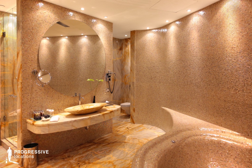 This high-end hotel bathroom exudes elegance with its seamless blend of warm-toned marble and intricate mosaic tiles. The floating marble vanity, featuring a sculpted stone basin, is complemented by a large circular mirror, enhancing the sense of space. Soft recessed lighting highlights the smooth, curved surfaces, while a built-in bathtub seamlessly integrates into the design. The overall aesthetic is a sophisticated balance of natural textures and modern luxury, making it an ideal setting for upscale hospitality, spa, or wellness-themed productions.