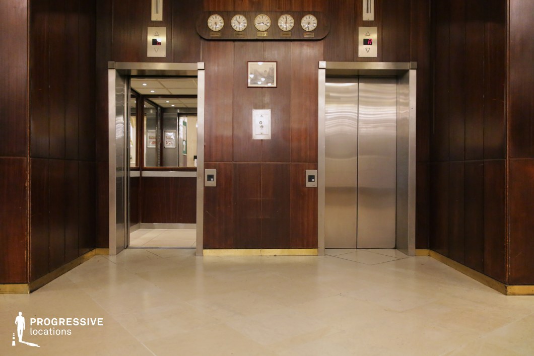 This elevator lobby features a vintage aesthetic with dark wooden paneling, brass details, and a set of dual elevators—one with a stainless steel door and the other open, revealing a mirrored interior. Above the elevators, a row of analog clocks displaying different time zones adds a retro charm. The polished stone flooring complements the warm wood tones, creating a sophisticated yet nostalgic ambiance. Ideal for classic hotel settings, period films, or upscale office locations requiring a timeless, elegant backdrop.