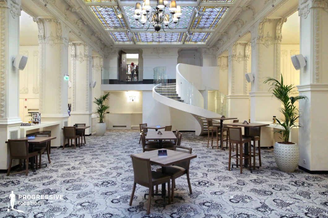 This sophisticated lobby blends classical architecture with modern elegance. The space is defined by towering, intricately carved columns supporting a high ceiling adorned with a stained-glass skylight. A dramatic white spiral staircase with a sleek glass railing leads to an upper level, adding a contemporary contrast to the historical detailing. The plush carpet, neutral-toned furniture, and lush greenery contribute to a refined yet inviting ambiance. Ideal for luxury settings, high-end events, or period film productions, this location offers a striking balance between old-world grandeur and modern design elements.