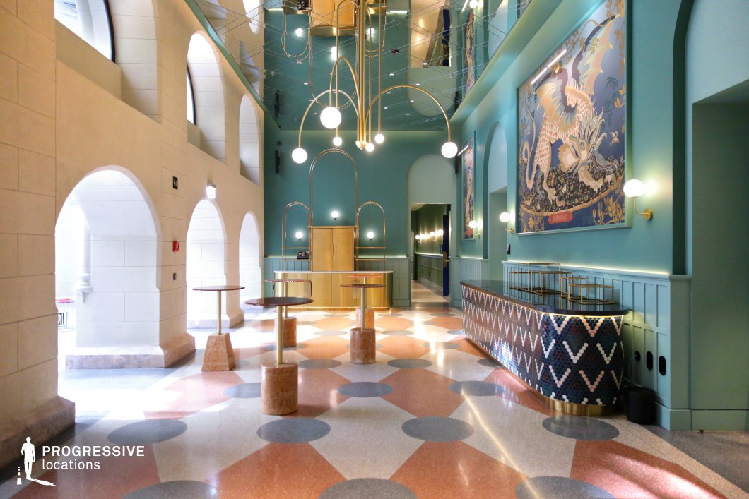This vibrant lobby blends historic and contemporary design elements, featuring grand arched openings, a high reflective ceiling, and a bold geometric terrazzo floor. The rich teal walls contrast elegantly with warm gold accents, including sculptural light fixtures and a brass reception desk. Oversized, whimsical artwork adds a playful yet sophisticated touch, while decorative mosaic tiling enhances the bar area. The combination of classical architecture and modern aesthetics makes this an ideal filming location for stylish dramas, artistic productions, or visually striking fantasy and surrealist narratives.