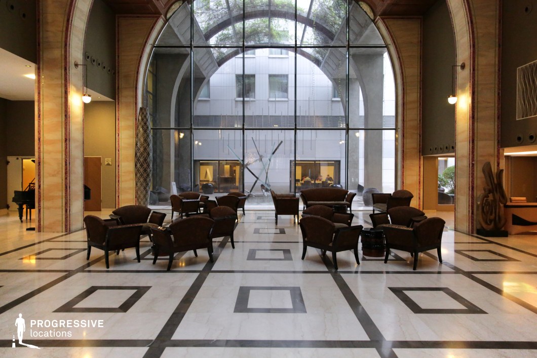 This expansive atrium lounge blends modern minimalism with classical grandeur, featuring towering arched windows that allow natural light to flood the space. The polished marble flooring, with geometric inlays, contrasts beautifully against the warm wood and soft neutral tones of the furniture. Sculptural elements and a dramatic glass façade add contemporary sophistication, while the arched architectural details evoke timeless elegance. This location is ideal for high-end dramas, corporate thrillers, or stylish espionage films, offering a refined and cinematic backdrop for power dynamics, negotiations, or moments of quiet contemplation.