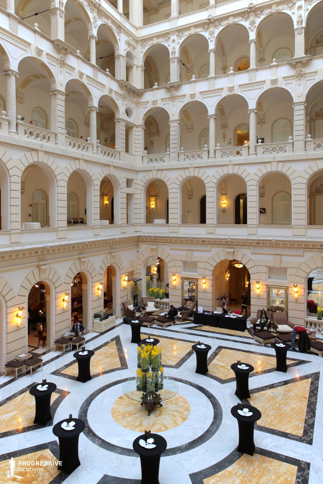 This magnificent atrium features towering arches, ornate balustrades, and a pristine white façade that exudes classical elegance. The multi-level design, accented with soft lighting and intricate moldings, enhances the sense of grandeur, while the polished marble flooring with bold geometric patterns adds a refined touch. The open central space, furnished with cocktail tables and elegant floral arrangements, is ideal for upscale gatherings or formal events. This location is perfect for period dramas, luxury settings, or high-society intrigue, offering a striking cinematic backdrop for pivotal scenes of power, elegance, or dramatic encounters.