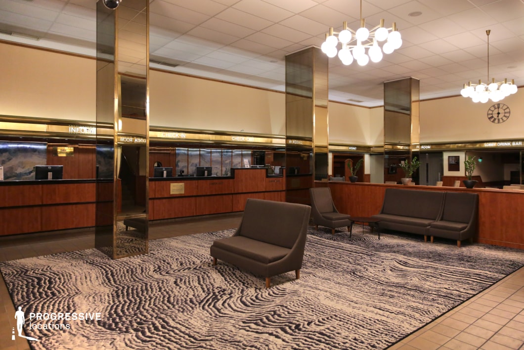 This hotel lobby showcases a vintage design with wood-paneled reception desks, gold-accented columns, and a ceiling lined with globe chandeliers. The patterned carpet in muted tones complements the dark gray seating, creating a mid-century atmosphere. Reflective gold surfaces and a large analog clock add to the nostalgic ambiance, reminiscent of Soviet-era architecture. The symmetrical layout, structured lighting, and functional seating arrangement make this space suitable for period productions, retro-themed settings, or classic hotel interiors.