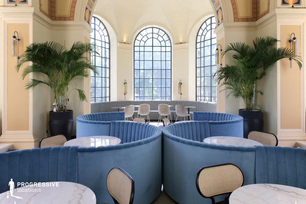 This refined lounge blends classical architecture with modern luxury, featuring grand arched windows that flood the space with natural light. The cream-colored walls with intricate moldings and subtle decorative accents enhance the sophisticated ambiance, complemented by deep blue velvet banquette seating arranged in a curved, symmetrical layout. Lush potted palms add a natural element, softening the structured design. The marble-topped tables and sleek contemporary chairs complete the upscale aesthetic, making this an ideal filming location for period dramas, high-society narratives, or romantic and stylish character interactions.