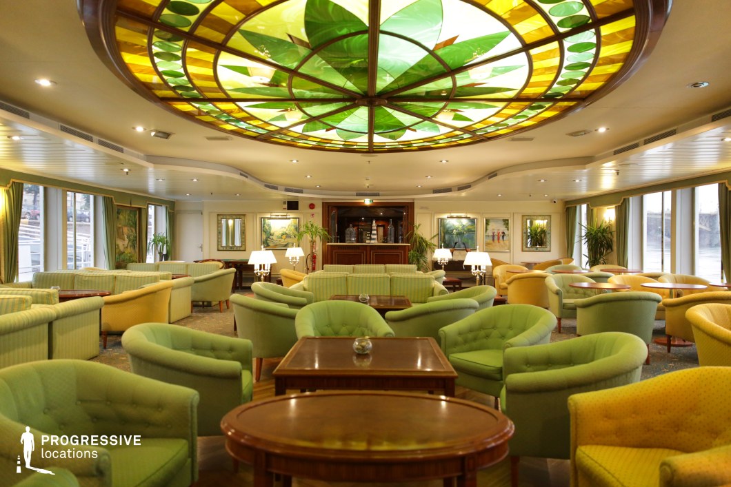 This vibrant and inviting lounge showcases a striking stained-glass ceiling with lush green and golden hues, serving as a captivating focal point. The plush armchairs and sofas in soft green and yellow tones create a relaxed yet refined atmosphere, complemented by polished wooden tables and classic decor elements. Large windows allow natural light to filter in, enhancing the airy and open feel of the space. The harmonious color palette and sophisticated furnishings make this an ideal setting for upscale social gatherings, casino or cruise ship scenes, and character-driven dramas, offering a stylish yet comfortable cinematic backdrop.