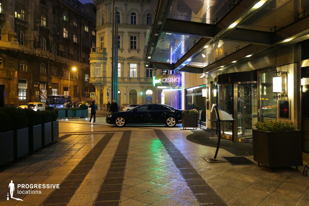 The entrance of a modern, high-end hotel is illuminated by artificial lighting, reflecting off the sleek glass facade and metal canopy. The setting is framed by a mix of contemporary and historic architecture, with a grand neoclassical building in the background contrasting with the modern urban landscape. A black luxury car is parked at the entrance, enhancing the scene’s exclusivity. Neon lights from a nearby casino create a vibrant city nightlife atmosphere. This location is ideal for filming high-stakes drama, sophisticated arrivals, or urban nightlife sequences.