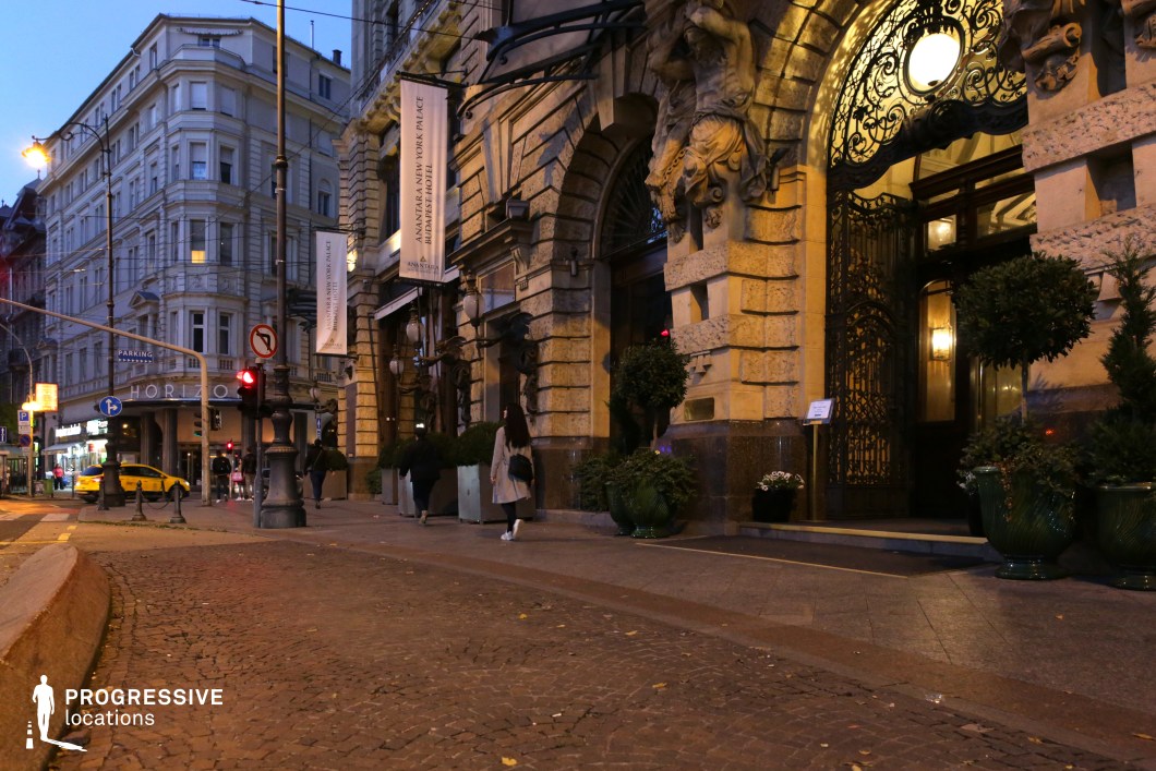 This ornate hotel entrance exudes timeless elegance, featuring an imposing stone façade adorned with sculptural reliefs and intricate wrought-iron gates. The warm glow of classic street lamps and interior lighting contrasts with the cool tones of the cobblestone street and historic architecture. The surrounding urban setting, with grand buildings and decorative elements, enhances the old-world sophistication of the location. This setting is ideal for noir thrillers, period dramas, or high-society intrigue, providing a dramatic and atmospheric backdrop for arrivals, clandestine meetings, or pivotal turning points in a story.