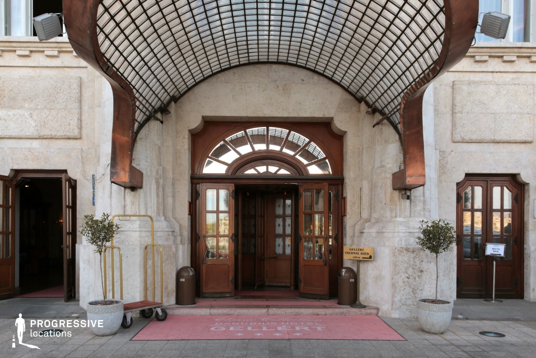 This striking entrance features a curved Art Nouveau-style glass and metal canopy, complementing the grand stone façade with intricate detailing. The wooden revolving door, framed by arched windows, adds an air of old-world sophistication. The combination of natural stone, warm wood, and ornamental metalwork creates a rich architectural contrast, while subtle decorative elements enhance the historic character. This location is ideal for period dramas, luxury hotel settings, or mystery thrillers, providing an elegant yet slightly enigmatic backdrop for scenes involving high society, intrigue, or clandestine meetings.