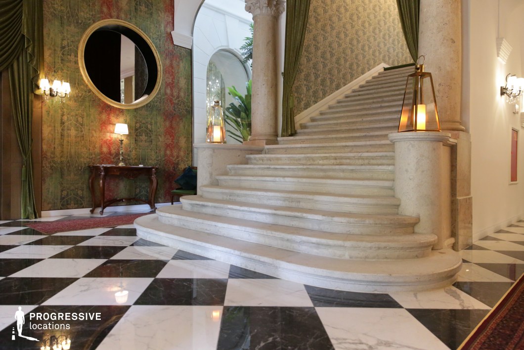 This luxurious interior showcases a grand, sweeping stone staircase framed by ornate columns and rich classical details. The black-and-white marble checkerboard flooring enhances the opulence of the space, reflecting the soft glow of the ambient lighting. A large circular mirror and deep green drapery add dramatic contrast to the warm tones of the textured wallpaper. The scene is further accentuated by antique-style furniture and decorative lanterns, making it an ideal setting for period dramas, high-end hospitality shoots, or sophisticated luxury branding.
