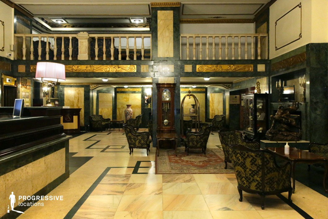 This stately hotel lobby exudes an old-world elegance with its rich marble surfaces in deep green and warm gold hues, complemented by intricate gilded moldings. The polished checkered floor and classical furniture, featuring dark wood and patterned upholstery, enhance the refined ambiance. A grand antique clock stands as a focal point, adding a timeless charm, while decorative elements such as sculptures and vintage lamps contribute to the atmosphere of sophistication. This setting is ideal for period dramas, noir thrillers, or mystery films, offering a luxurious and slightly enigmatic backdrop for intrigue and high-stakes encounters.
