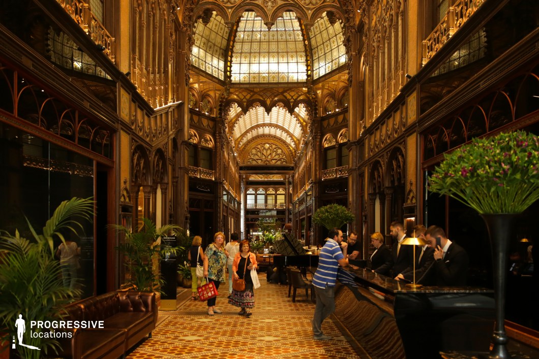 This opulent hotel lobby showcases intricate neo-Gothic and Art Nouveau architectural elements, featuring grand archways, elaborate gold-accented detailing, and a breathtaking stained-glass ceiling. The space is lined with dark wood paneling, decorative ironwork, and mirrored surfaces that enhance its luxurious and historic ambiance. The patterned tile flooring adds to the grandeur, while lush greenery softens the formal aesthetic. The symmetrical composition, combined with warm golden lighting, makes this an ideal setting for period dramas, high-society intrigue, or noir-inspired mystery films, exuding an air of sophistication and timeless elegance.