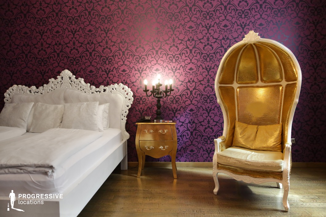 This lavish bedroom blends classic European opulence with dramatic color contrasts. A white, intricately carved bed frame stands against deep purple damask-patterned wallpaper, exuding aristocratic elegance. A striking golden high-back armchair with a glossy reptilian texture complements the ornate aesthetic, while a gilded nightstand and black candelabra-style lamp enhance the regal ambiance. The wooden flooring balances the richness of the decor. This setting is ideal for period dramas, fantasy productions, or eccentric, high-fashion editorial shoots requiring a bold and extravagant backdrop.