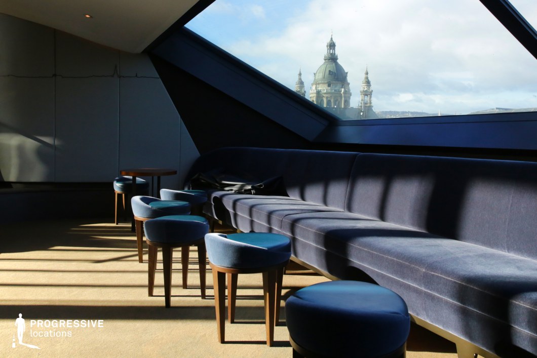 This sleek and contemporary lounge features deep blue velvet seating and matching stools, arranged beneath angular black-framed windows that frame a striking view of a historic domed building. The geometric architecture and soft natural light create an intimate yet futuristic atmosphere, with the interplay of shadows adding depth and contrast. The minimalist interior design, with clean lines and subtle textures, makes this an ideal filming location for sophisticated dramas, psychological thrillers, or high-end lifestyle productions that emphasize elegance with a modern edge.