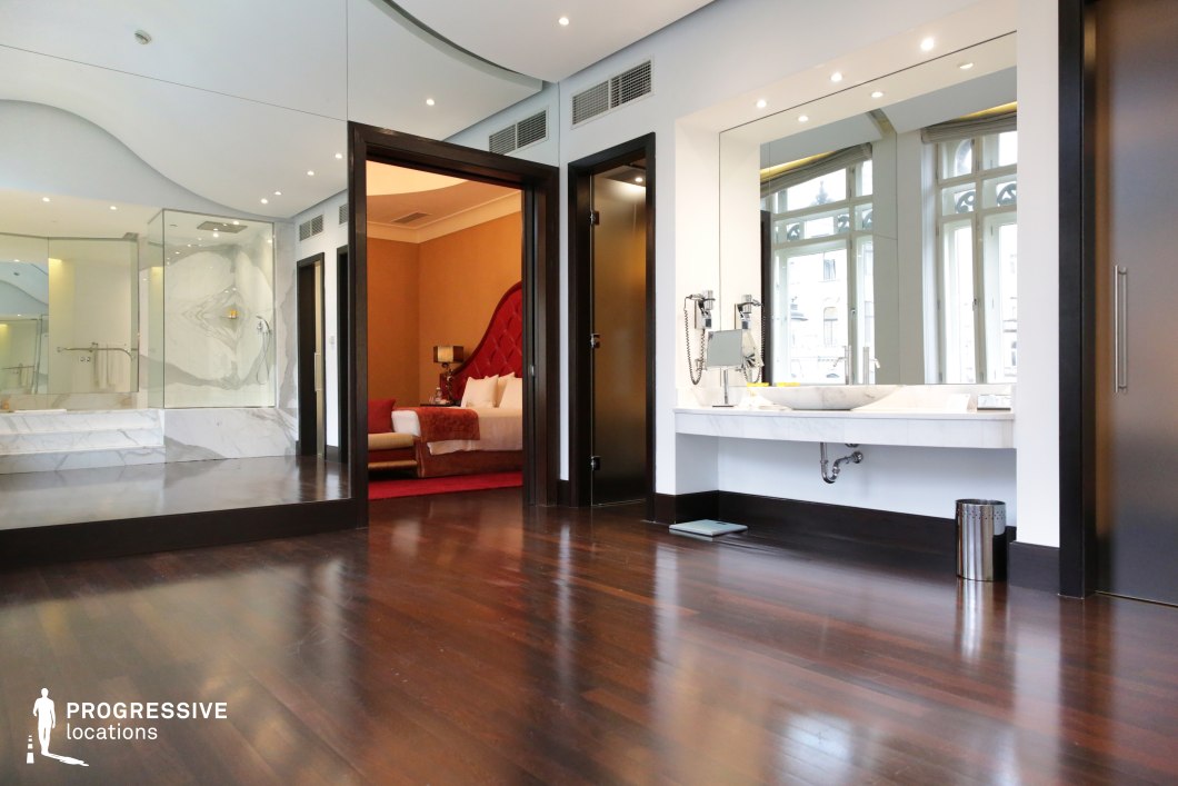 This high-end hotel suite features a sleek combination of dark wood flooring, marble accents, and expansive mirrors that enhance the sense of space and luxury. The open-concept layout seamlessly connects the bedroom, bathroom, and vanity area, with a striking red upholstered headboard adding a bold contrast to the neutral tones. Large arched windows allow natural light to highlight the polished surfaces and refined architectural details. The sophisticated yet inviting ambiance makes this an ideal filming location for dramas, romance, or psychological thrillers requiring an elegant and modern setting.