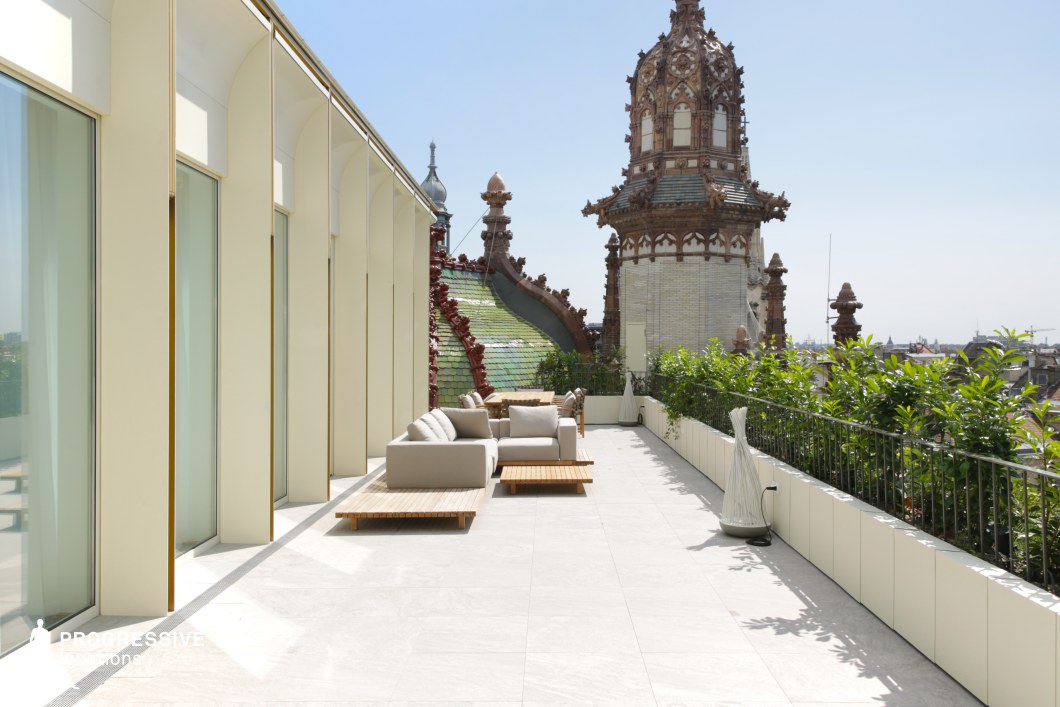 This elegant rooftop terrace seamlessly blends modern minimalism with historical grandeur, featuring sleek white flooring, floor-to-ceiling glass windows, and contemporary outdoor seating. The lush greenery along the railing softens the structured design while framing the ornate architectural details of the adjacent historic building, including intricate towers and multicolored roof tiles. The contrast between the clean, linear facade and the rich textures of the historic backdrop creates a visually striking setting, ideal for filming high-end lifestyle scenes, romance, or sophisticated drama requiring a blend of modern luxury and old-world charm.