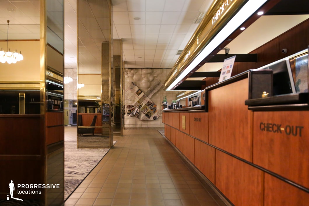 This hotel reception area exudes a mid-century elegance with its wood-paneled check-in counters, gold-accented signage, and reflective mirror columns that enhance the sense of depth and symmetry. The tiled flooring and structured ceiling design contribute to a retro yet sophisticated ambiance, while the large decorative wall piece in the background adds a unique visual focal point. The warm lighting and combination of polished and textured surfaces create a setting that is ideal for filming period dramas, noir thrillers, or political intrigue films, emphasizing luxury with a subtle undertone of nostalgia.