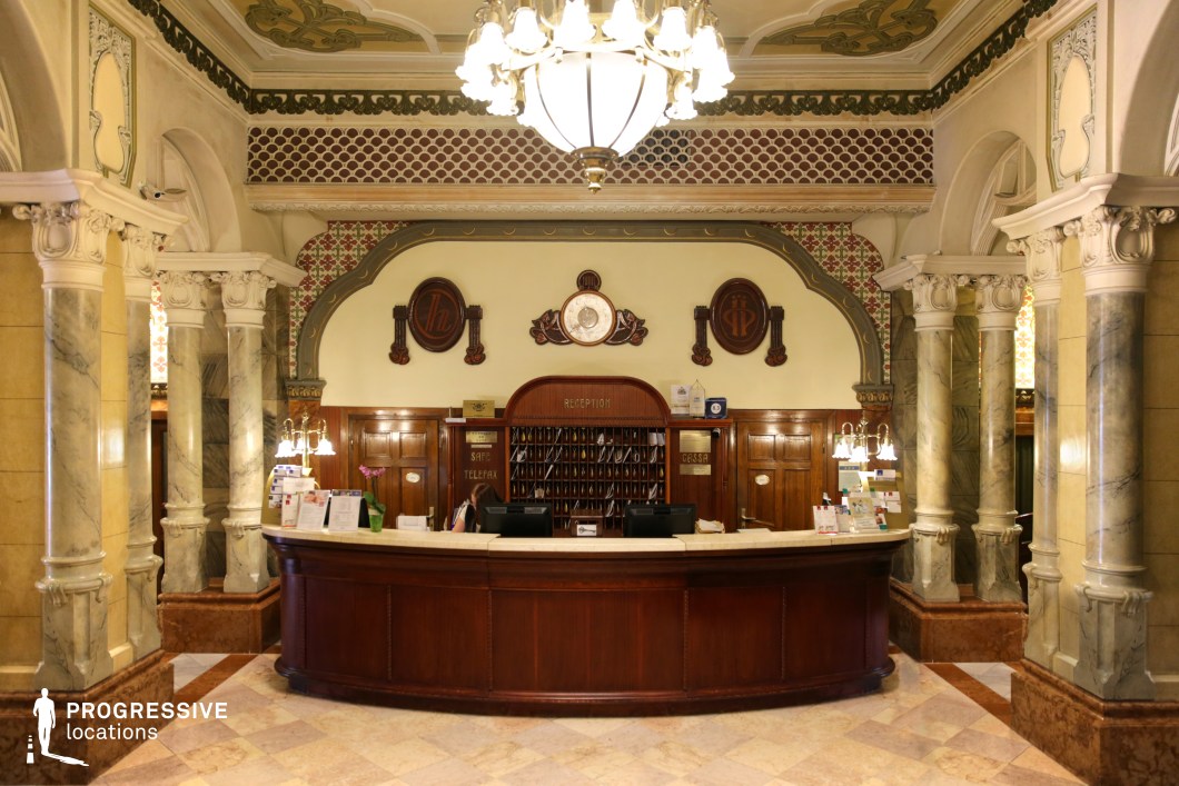 This grand hotel reception area exudes elegance with its richly detailed Art Nouveau and classical influences. The curved wooden desk, framed by decorative marble columns, creates a striking focal point, while the intricately patterned ceiling and wall motifs add historical charm. Soft, ambient lighting from vintage chandeliers enhances the warm tones of the polished wood paneling and luxurious marble flooring. This setting is ideal for period dramas, mystery thrillers, or historical romance films, providing a refined and visually intricate backdrop that evokes sophistication and old-world hospitality.
