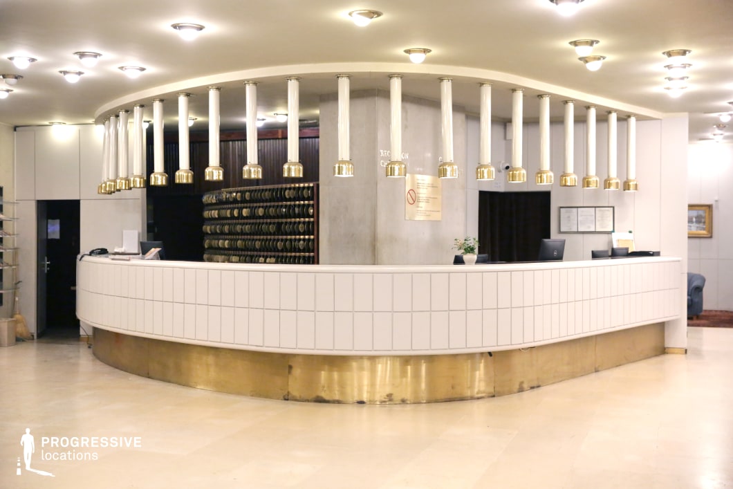 This curved hotel reception desk showcases a mid-century modern aesthetic with white tile paneling and a brass base, evoking a sense of retro luxury. Above the counter, a series of suspended white and gold cylindrical light fixtures add a striking visual element, enhancing the vintage appeal. The key organizer wall behind the counter, paired with dark wood paneling, reinforces the classic hospitality atmosphere. The polished stone flooring reflects the soft ambient lighting, contributing to a warm yet formal ambiance. This setting is ideal for film or photography projects requiring a nostalgic or Soviet-era inspired hotel interior.