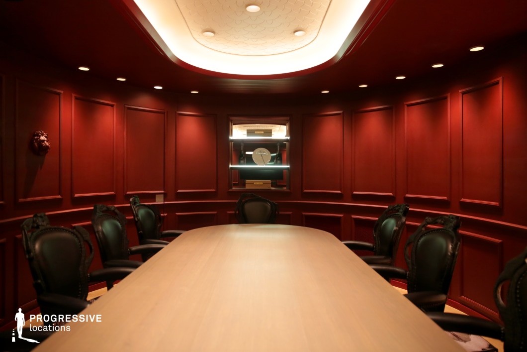 This intimate meeting space exudes sophistication with its deep red paneling, curved walls, and ambient recessed lighting. The centerpiece is a sleek wooden conference table surrounded by black leather chairs with ornate detailing, reinforcing a classic and exclusive aesthetic. A softly illuminated ceiling feature adds depth, while a built-in display case at the far end enhances the refined atmosphere. The dramatic yet elegant setting makes this an ideal filming location for political thrillers, corporate dramas, or high-stakes negotiation scenes requiring a sense of secrecy and power.