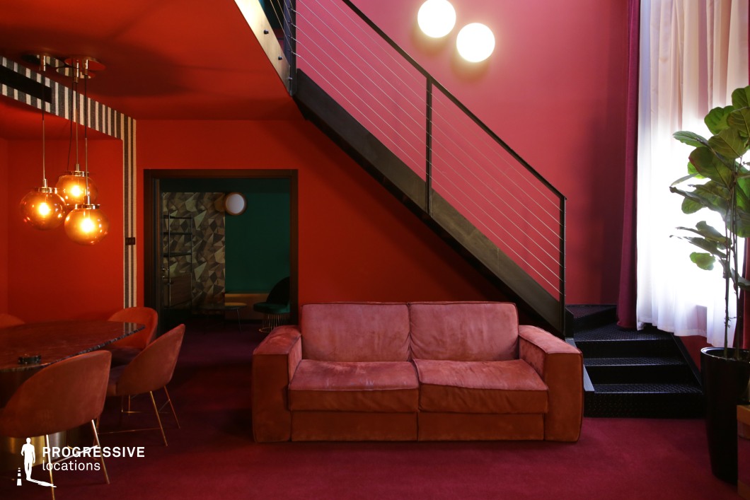 This striking lounge space features deep red walls and carpeting, complemented by a plush matching sofa and bold geometric accents. A sleek black metal staircase adds contrast, while warm-toned pendant lights enhance the vintage-inspired aesthetic. The open doorway reveals an adjacent room with a mix of green and geometric wall textures, adding depth and visual intrigue. The combination of rich colors, eclectic design, and layered spaces makes this an ideal filming location for stylish period pieces, artistic dramas, or retro-inspired thrillers.