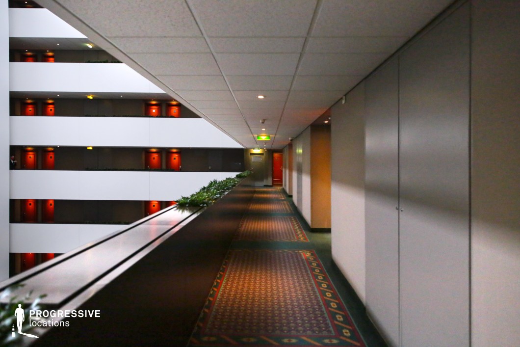 This modern hotel corridor overlooks a multi-story atrium, featuring a sleek railing lined with greenery. The patterned carpet with green and red tones complements the deep red doors on the opposite balconies, creating a visually striking contrast against the white walls. The repetitive architectural elements and long, linear perspective add a sense of depth and symmetry. The blend of contemporary and retro aesthetics, combined with the moody lighting, makes this an ideal setting for filming thrillers, corporate dramas, or psychological suspense films that require a structured yet subtly unsettling atmosphere.