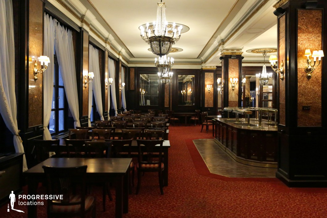 This refined dining hall showcases an opulent blend of dark wood paneling, gilded detailing, and deep red carpeting, evoking the grandeur of early 20th-century establishments. Large windows draped with sheer white curtains create an elegant contrast against the rich interior, while ornate chandeliers and classic wall sconces provide a warm, ambient glow. The polished wooden tables and plush seating contribute to the sophisticated atmosphere. This setting is ideal for period dramas, noir thrillers, or high-society intrigue, offering a cinematic backdrop for elegant gatherings, clandestine meetings, or dramatic confrontations.