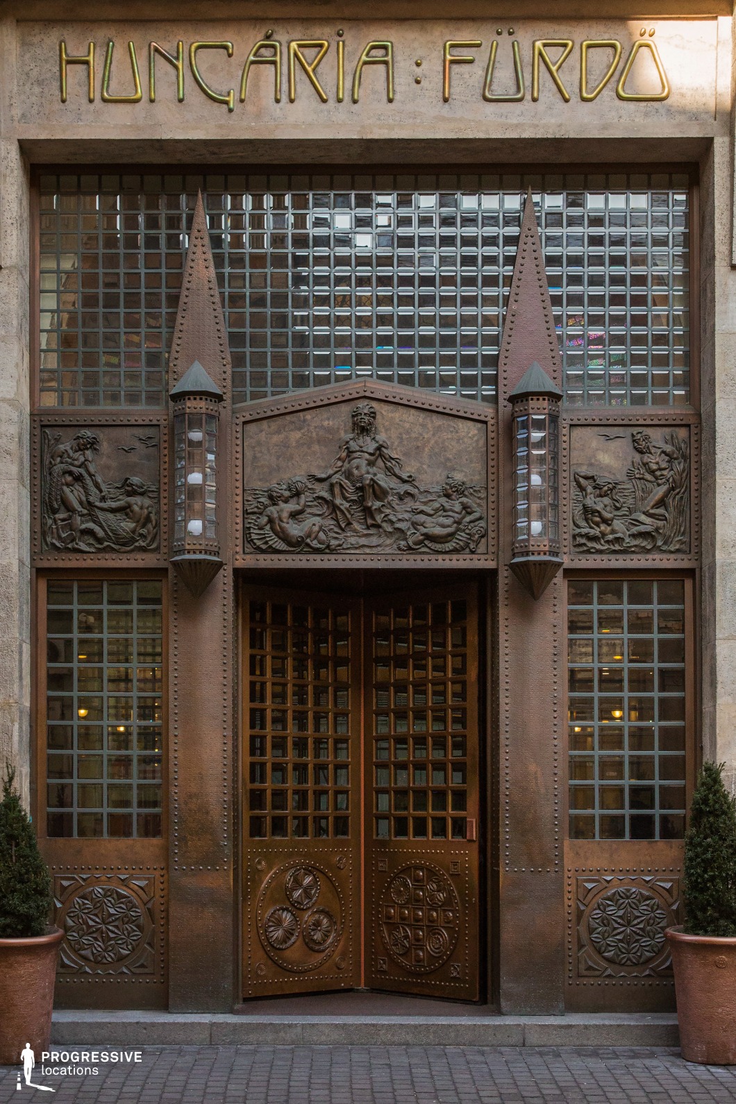 This grand entrance showcases a striking Art Nouveau design, featuring an intricately detailed bronze doorway adorned with sculpted reliefs of mythological figures and decorative geometric patterns. Flanked by tall, grid-like glass panels, the entrance exudes a sense of historic elegance and craftsmanship. The riveted metalwork and pointed architectural embellishments enhance the structure’s imposing presence, while the soft glow from within adds depth to the composition. This setting is ideal for period dramas, noir thrillers, or mystery films, offering a cinematic backdrop that suggests exclusivity, hidden histories, and an air of intrigue.