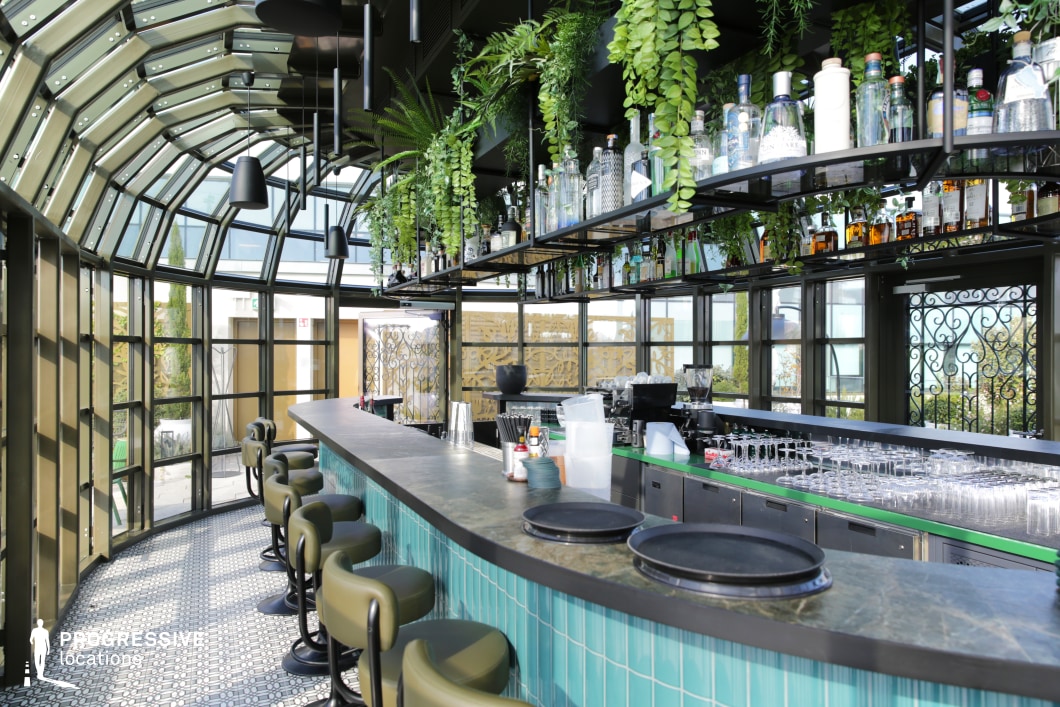 This sophisticated rooftop bar features a striking arched glass ceiling and walls, allowing ample natural light to highlight the vibrant green and turquoise color scheme. Suspended greenery and industrial black metal shelving filled with bottles create a lush, modern aesthetic, while the curved bar lined with plush olive-green stools enhances the intimate yet stylish atmosphere. The intricate tile flooring and wrought-iron accents add a touch of elegance. This setting is ideal for filming upscale social scenes, nightlife sequences, or contemporary dramas requiring a chic, cosmopolitan backdrop.