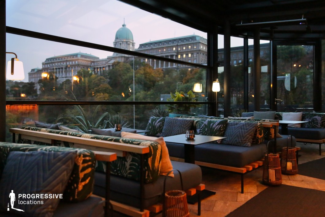 This stylish rooftop lounge blends contemporary design with a breathtaking historic backdrop, featuring floor-to-ceiling glass windows overlooking a grand domed castle. The interior combines dark wood, plush seating in deep blue tones, and botanical-patterned cushions, creating a cozy yet sophisticated atmosphere. Soft lighting enhances the ambiance, while woven lanterns and greenery add texture and warmth. The seamless integration of modern elegance and historic scenery makes this an ideal filming location for romance, high-society dramas, or stylish city nightlife scenes.